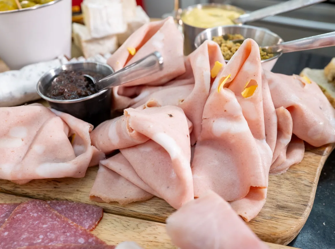 Folds of pale pink mortadella on a wooden board beside small pots of relish